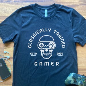 May include: Black t-shirt with a white skull graphic wearing a retro video game controller as glasses. The text reads "Classically Trained Gamer" with the years "ESTD 1990" underneath.
