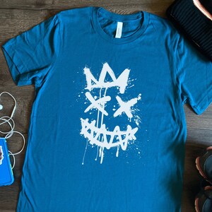 Puede incluir: Una camiseta azul turquesa con un gráfico blanco de una cara estilizada con una corona y pintura salpicada.