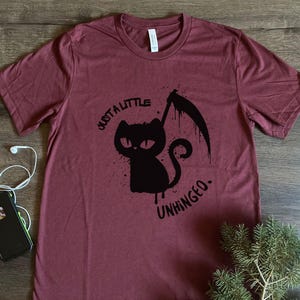 May include: Burgundy t-shirt with a black graphic of a cat holding a scythe and the text "Just a little unhinged." The shirt is made of a soft material and has a crew neck.