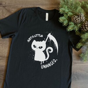 May include: Black t-shirt with a white graphic of a cat holding a scythe. The text "just a little unhinged" is printed around the cat. The shirt is made of a soft material.