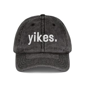 May include: A faded black baseball cap featuring the word "yikes." in white lettering. The cap has a curved peak and an adjustable fabric strap at the back.