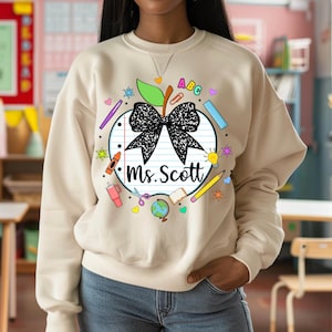 Puede incluir: Una sudadera beige con un gráfico de un lazo negro sobre una manzana roja con el texto "ABC" y "Ms. Scott" rodeado de útiles escolares coloridos.