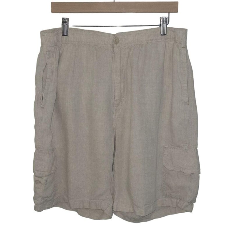 May include: Tan linen cargo shorts hanging on a wooden hanger. The shorts have an elastic waistband, a button closure, and two side pockets. There are two additional cargo pockets on the legs. The shorts are made of a lightweight, breathable fabric.