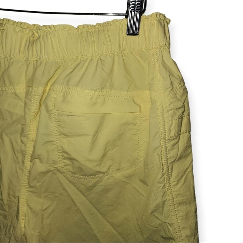 May include: Close-up of yellow shorts with an elasticated waistband and a single back pocket. The shorts are made of a lightweight, slightly creased fabric. The shorts are hanging on a metal hanger.