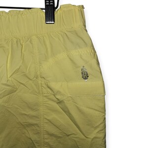 May include: Pale yellow athletic shorts with an elasticated waistband and a small pocket. The shorts have a subtle sheen and a small logo on the pocket. The fabric appears lightweight, suitable for exercise or casual wear.