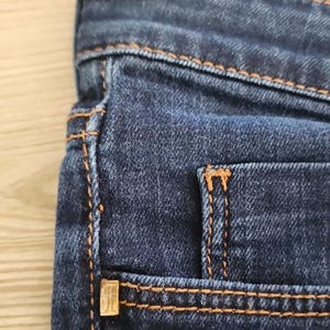 May include: Close-up of dark blue denim jeans. The jeans have orange stitching along the seams and around the pockets. A small, gold-coloured metal detail is visible on the pocket.