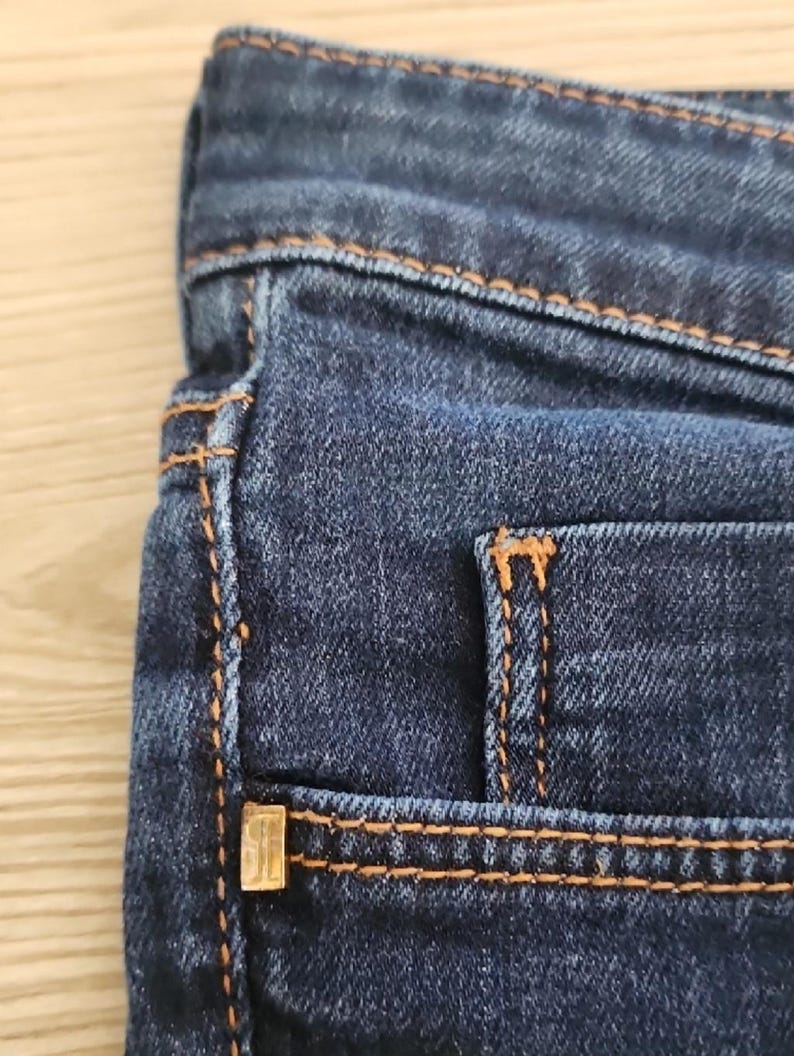 May include: Close-up of dark blue denim jeans. The jeans have orange stitching along the seams and around the pockets. A small, gold-coloured metal detail is visible on the pocket.