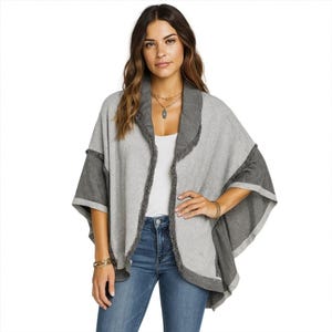 Op de afbeelding: Een grijze en donkergrijze open poncho met een gefranjerde kraag en mouwranden. De poncho is gemaakt van een zacht materiaal en heeft een relaxte pasvorm. Het model draagt een blauwe spijkerbroek en een witte top.
