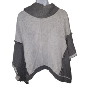 Op de afbeelding: Een grijze en houtskoolkleurige poncho-achtige top met een col. De mouwen en zoom zijn houtskool, met een lichtere grijze body. De naden hebben een rauwe, onafgewerkte rand. De top wordt getoond op een witte mannequin.
