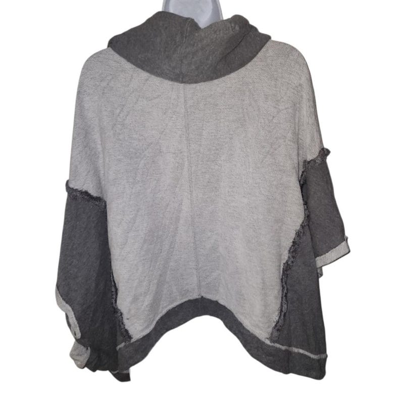 Op de afbeelding: Een grijze en houtskoolkleurige poncho-achtige top met een col. De mouwen en zoom zijn houtskool, met een lichtere grijze body. De naden hebben een rauwe, onafgewerkte rand. De top wordt getoond op een witte mannequin.