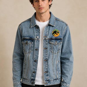 May include: Light blue denim jacket with a classic collar, button-down front, and chest pockets. Features a colourful circular patch on the upper left chest with a dinosaur graphic. The jacket is worn over a white t-shirt.