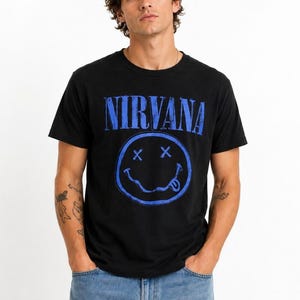 Puede incluir: Una camiseta negra de manga corta con la palabra NIRVANA en una fuente azul desgastada sobre un gráfico azul de una cara sonriente con ojos en forma de X y la lengua fuera. Se combina con unos vaqueros de color azul claro.