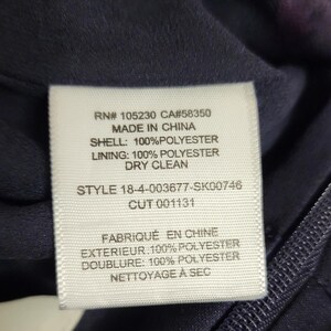 May include: Close-up of a white clothing tag with black text. The tag includes the text "RN# 105230 CA#58350 MADE IN CHINA SHELL: 100% POLYESTER LINING: 100% POLYESTER DRY CLEAN STYLE 18-4-003677-SK00746 CUT 001131". The tag is on a dark blue fabric.