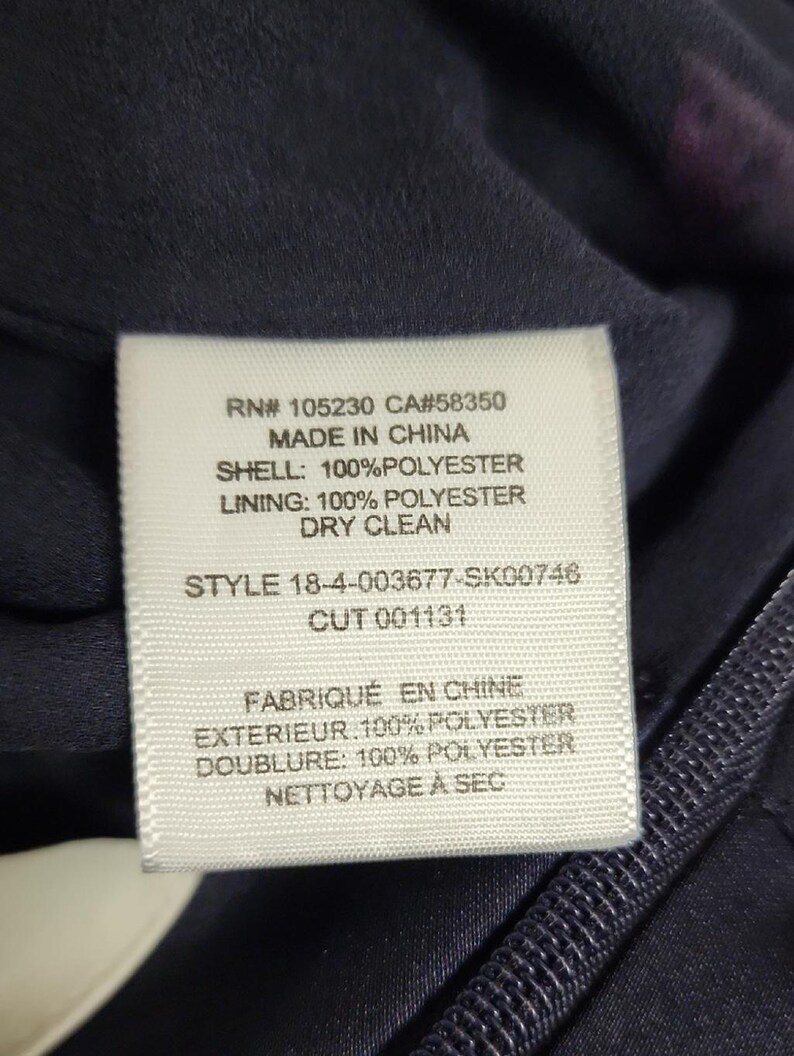 May include: Close-up of a white clothing tag with black text. The tag includes the text "RN# 105230 CA#58350 MADE IN CHINA SHELL: 100% POLYESTER LINING: 100% POLYESTER DRY CLEAN STYLE 18-4-003677-SK00746 CUT 001131". The tag is on a dark blue fabric.