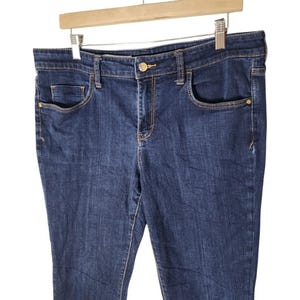 May include: Dark wash denim jeans with a classic five-pocket design. The jeans are hanging on a wooden hanger. Features include a button closure, belt loops, and gold-toned hardware accents on the pockets.