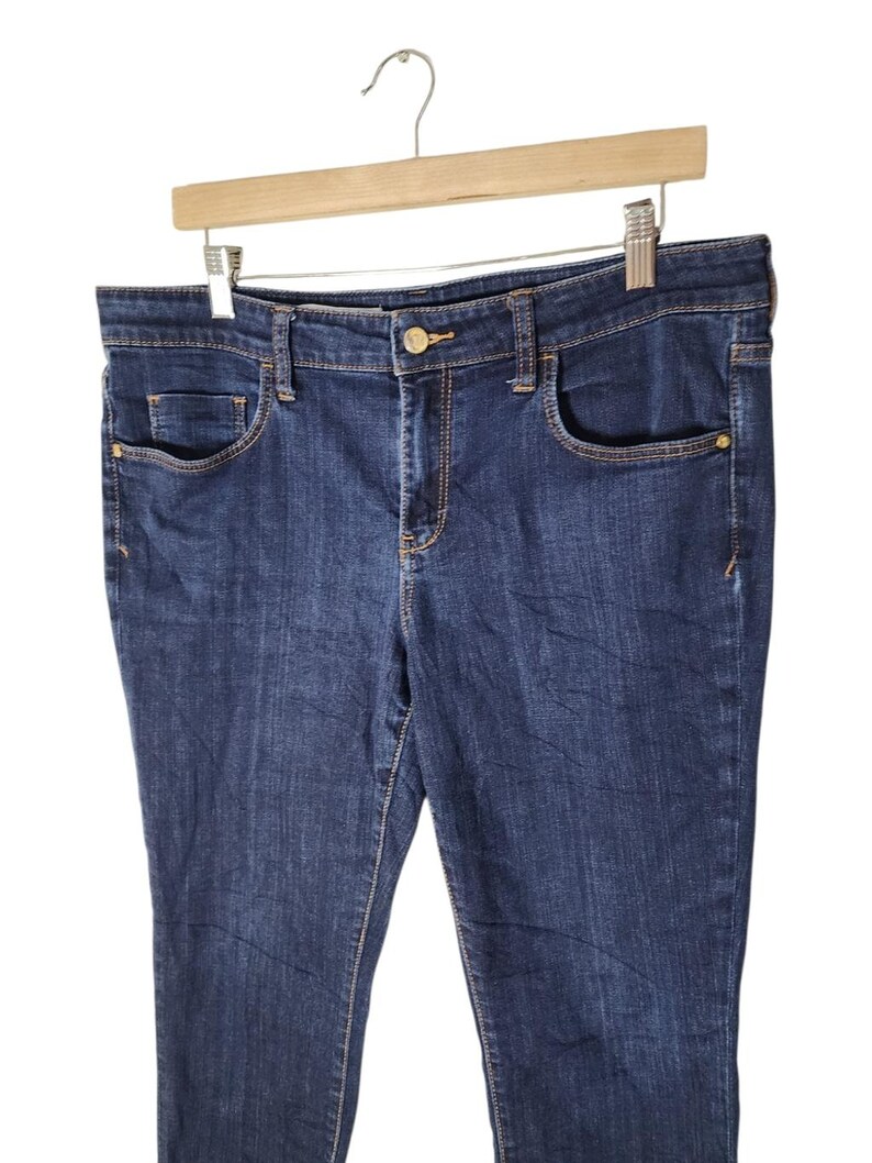 May include: Dark wash denim jeans with a classic five-pocket design. The jeans are hanging on a wooden hanger. Features include a button closure, belt loops, and gold-toned hardware accents on the pockets.