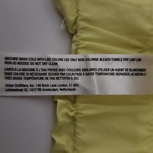 May include: A white label with black text providing garment care instructions. The label includes washing, bleaching, drying, and ironing instructions. The label also lists the Urban Outfitters address in London and Amsterdam.