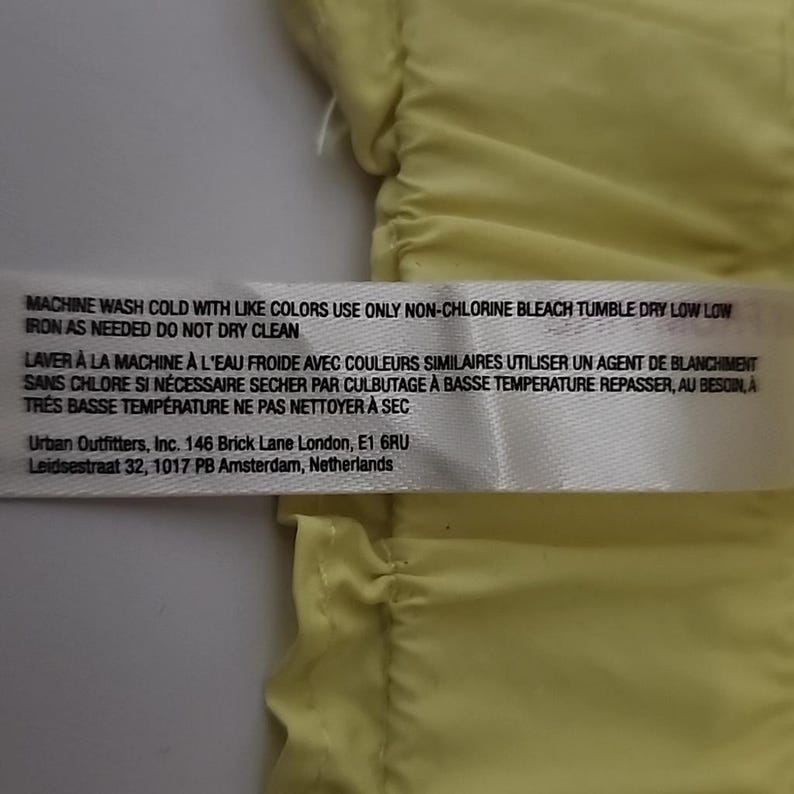 May include: A white label with black text providing garment care instructions. The label includes washing, bleaching, drying, and ironing instructions. The label also lists the Urban Outfitters address in London and Amsterdam.