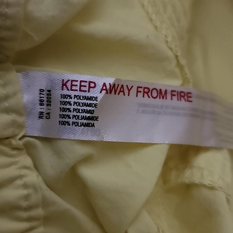 May include: A close-up shot of a white label with the red text "KEEP AWAY FROM FIRE". The label also states "100% POLYAMIDE" in white. The label is attached to a pale yellow fabric.