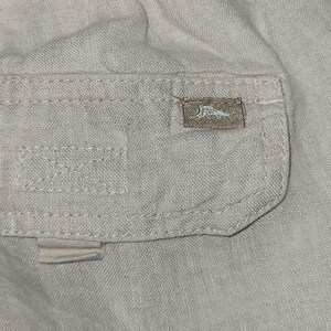 May include: Close-up of a light beige fabric with a small pocket. The pocket has a rectangular label with a logo. The fabric has visible stitching and a textured appearance. The overall aesthetic is casual and relaxed.