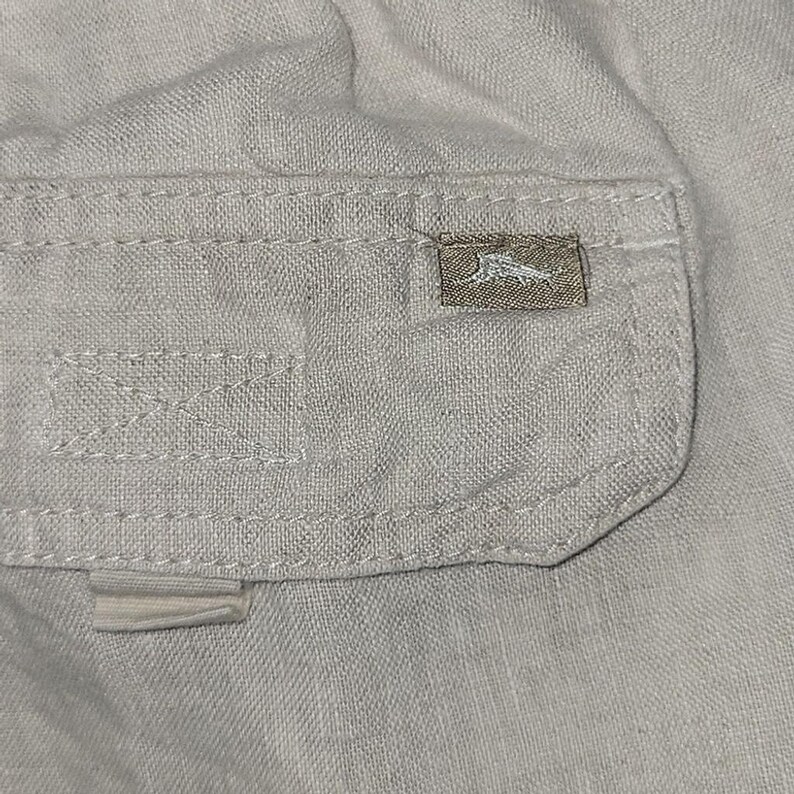 May include: Close-up of a light beige fabric with a small pocket. The pocket has a rectangular label with a logo. The fabric has visible stitching and a textured appearance. The overall aesthetic is casual and relaxed.