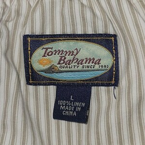 May include: Close-up of a garment tag with the Tommy Bahama logo, featuring a beach scene. The tag also includes the text "QUALITY SINCE 1993", size "L", and "100% LINEN MADE IN CHINA". The background is a striped fabric.