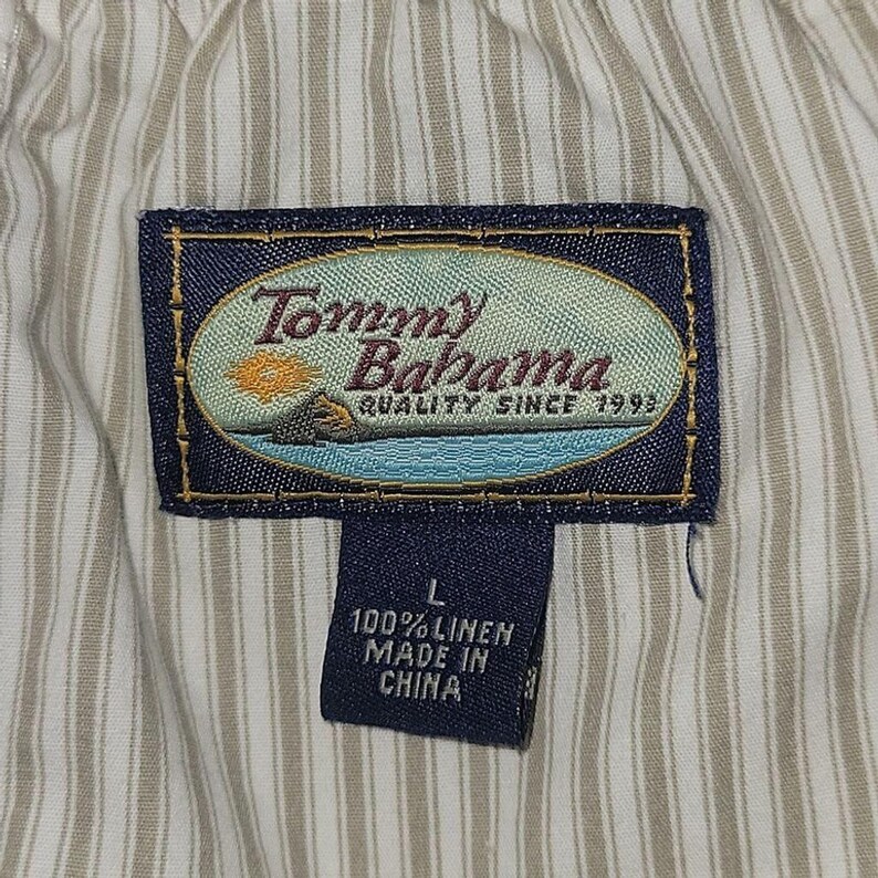 May include: Close-up of a garment tag with the Tommy Bahama logo, featuring a beach scene. The tag also includes the text "QUALITY SINCE 1993", size "L", and "100% LINEN MADE IN CHINA". The background is a striped fabric.