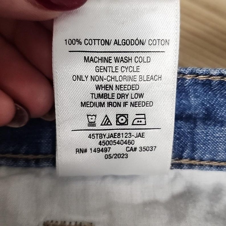 May include: A white fabric care label attached to blue denim fabric. The text reads 100% COTTON, MACHINE WASH COLD, GENTLE CYCLE, ONLY NON-CHLORINE BLEACH WHEN NEEDED, TUMBLE DRY LOW, MEDIUM IRON IF NEEDED, 45TBYJAE8123-JAE, 4500540460, RN# 149497, CA# 35037, 05/2023.