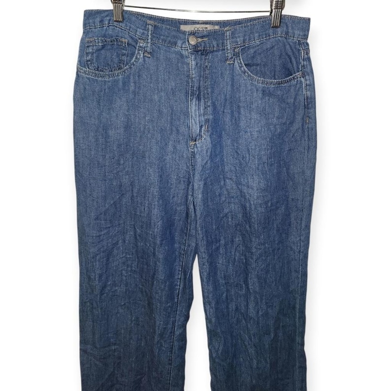 May include: Medium blue denim jeans featuring a high-waisted design with a classic five-pocket construction, a metal button closure, a zip fly, and belt loops along the waistband. The fabric has a consistent medium wash finish throughout.