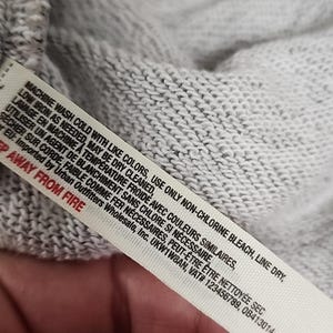 Op de afbeelding: Close-up van een grijze gebreide stof met een wit waslabel. Het label bevat wasvoorschriften in het Engels en Frans, zoals "Machine wash cold" en "Line dry". Op het label staat ook "Keep away from fire".