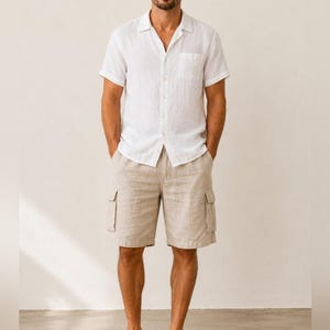 May include: A white short-sleeve shirt with a front pocket and beige cargo shorts. The shorts have two side pockets. The person is wearing brown sandals. The outfit is casual and suitable for warm weather.