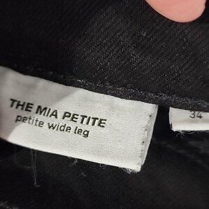 May include: Close-up of a black garment with a white tag. The tag reads "THE MIA PETITE petite wide leg." A smaller tag with the number "34" is also visible. The fabric appears to be a dark denim or similar material.