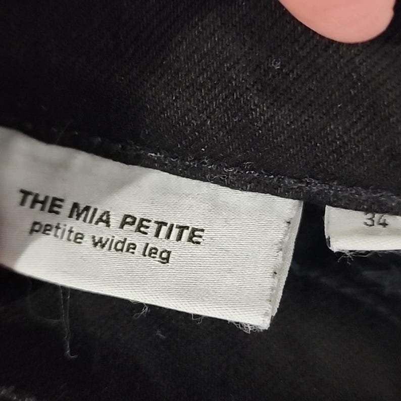 May include: Close-up of a black garment with a white tag. The tag reads "THE MIA PETITE petite wide leg." A smaller tag with the number "34" is also visible. The fabric appears to be a dark denim or similar material.