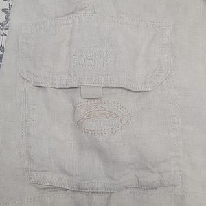 May include: A close-up of a light beige linen cargo pocket with a stitched fish design and the word "SPORT" embroidered on the pocket flap.