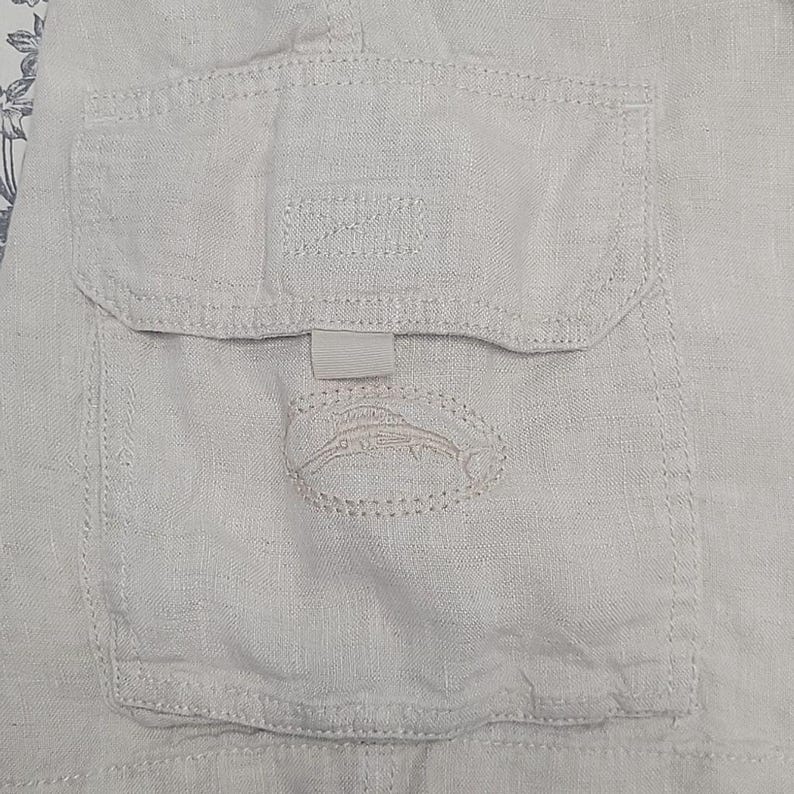 May include: A close-up of a light beige linen cargo pocket with a stitched fish design and the word "SPORT" embroidered on the pocket flap.