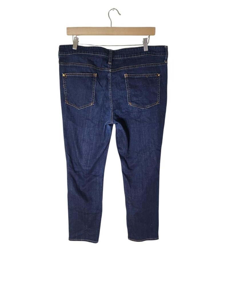 May include: Dark wash denim jeans with a classic five-pocket design, suspended from a wooden hanger. The jeans have a straight leg cut and gold-coloured hardware on the back pockets.