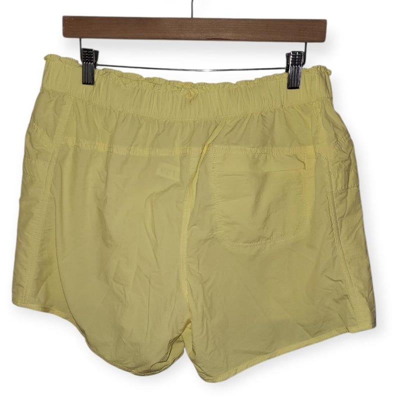May include: Pale yellow athletic shorts with an elasticated waistband and a single back pocket. The shorts are made of a lightweight, slightly crinkled fabric and hang from a wooden hanger.