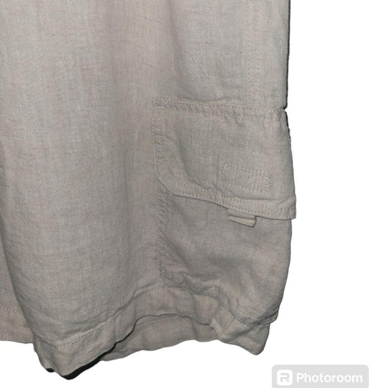 May include: A pair of light beige linen shorts with a large cargo pocket on the right side. The pocket has a flap with a button closure.
