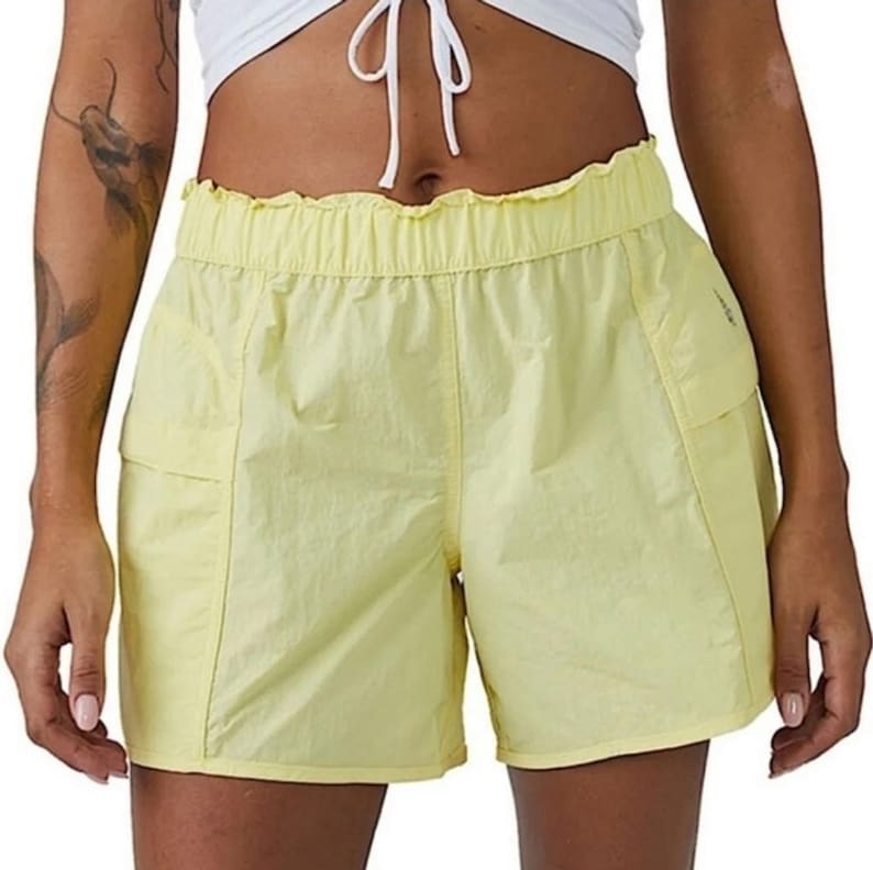 May include: Pale yellow shorts with a frilled waistband and two side pockets. The shorts are made of a lightweight, slightly sheer fabric. The shorts are a casual style.