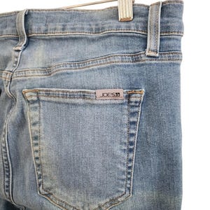 May include: Close-up of light blue denim jeans. The back pocket features a small rectangular tag with the brand name "JOE'S" in a dark color. The jeans are hanging on a metal hanger.