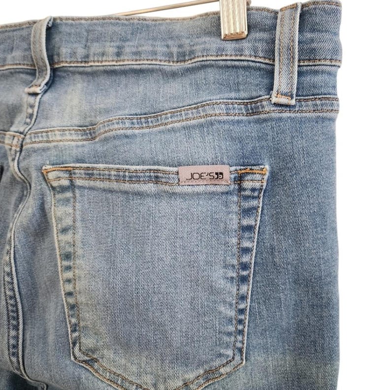 May include: Close-up of light blue denim jeans. The back pocket features a small rectangular tag with the brand name "JOE'S" in a dark color. The jeans are hanging on a metal hanger.
