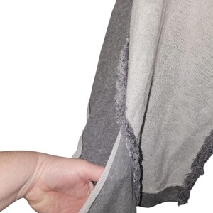 Op de afbeelding: Een grijze en lichtgrijze sweatshirt met een gerafeld randontwerp. De sweatshirt heeft een splitnaad aan de zijkant en een zak. De stof lijkt een zacht, comfortabel materiaal te zijn.