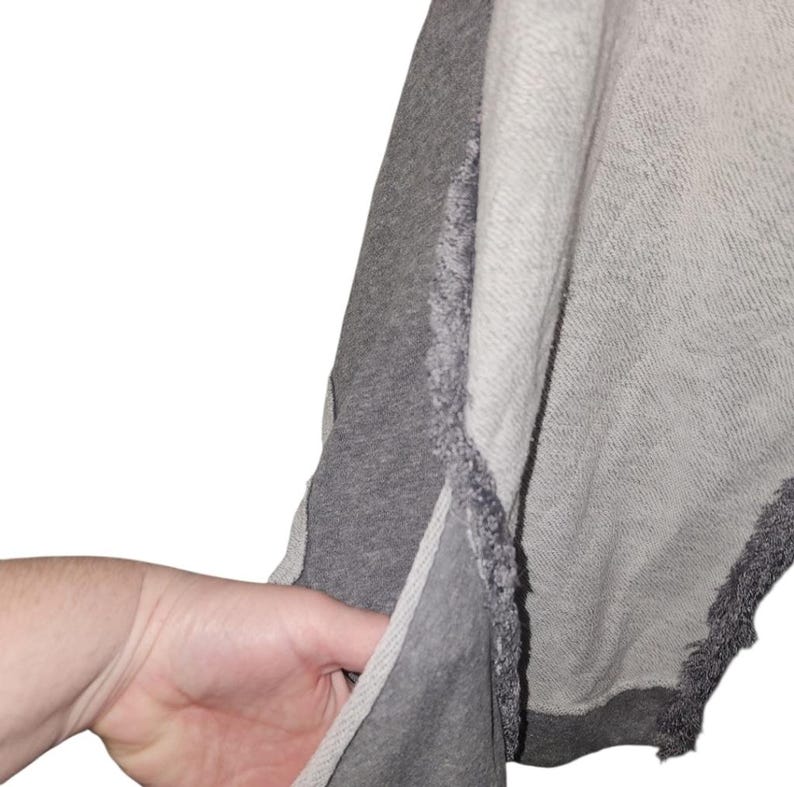 Op de afbeelding: Een grijze en lichtgrijze sweatshirt met een gerafeld randontwerp. De sweatshirt heeft een splitnaad aan de zijkant en een zak. De stof lijkt een zacht, comfortabel materiaal te zijn.
