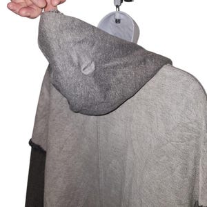 Op de afbeelding: Een grijze poncho met capuchon en een tweekleurig ontwerp. De capuchon is donkerder grijs dan de body van de poncho. De mouwen zijn donkerder grijs met een zwarte bies. De poncho hangt aan een witte hanger.