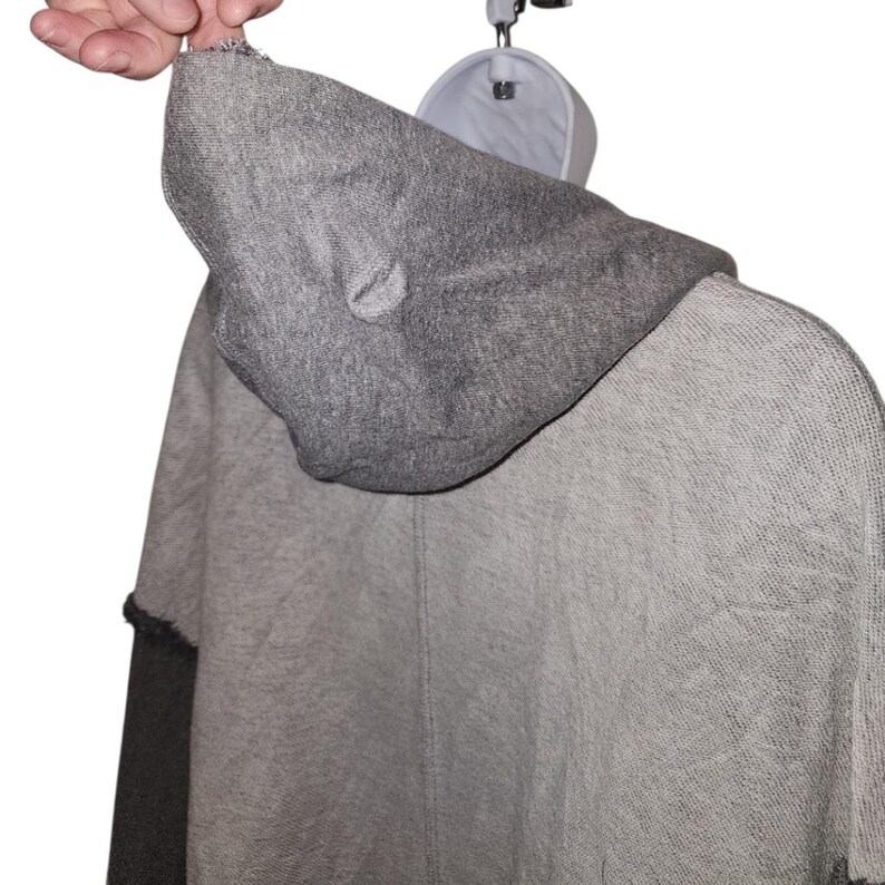 Op de afbeelding: Een grijze poncho met capuchon en een tweekleurig ontwerp. De capuchon is donkerder grijs dan de body van de poncho. De mouwen zijn donkerder grijs met een zwarte bies. De poncho hangt aan een witte hanger.