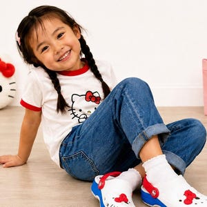 Peut inclure: Un t-shirt blanc à col rouge avec un motif Hello Kitty, un jean bleu, des chaussettes blanches et des sabots blancs ornés de détails rouges Hello Kitty, d'une semelle bleue et d'une bride arrière rouge. La chaussure présente des perforations.