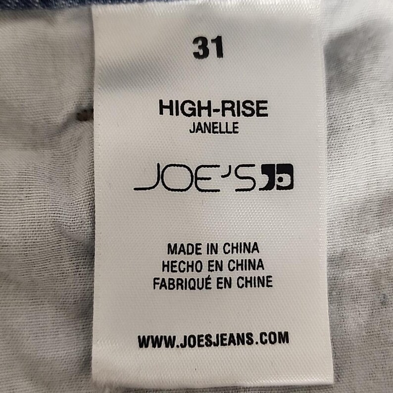May include: A white clothing label featuring black text that reads 31, HIGH-RISE, JANELLE, JOE'S, MADE IN CHINA, HECHO EN CHINA, FABRIQU&Eacute; EN CHINE, and WWW.JOESJEANS.COM, sewn onto the inside of blue denim jeans with a clear woven fabric texture.