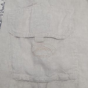 May include: Close-up of a light beige linen fabric with a pocket. The pocket has a flap and a small, stitched emblem of a fish. The fabric has visible texture and stitching details.