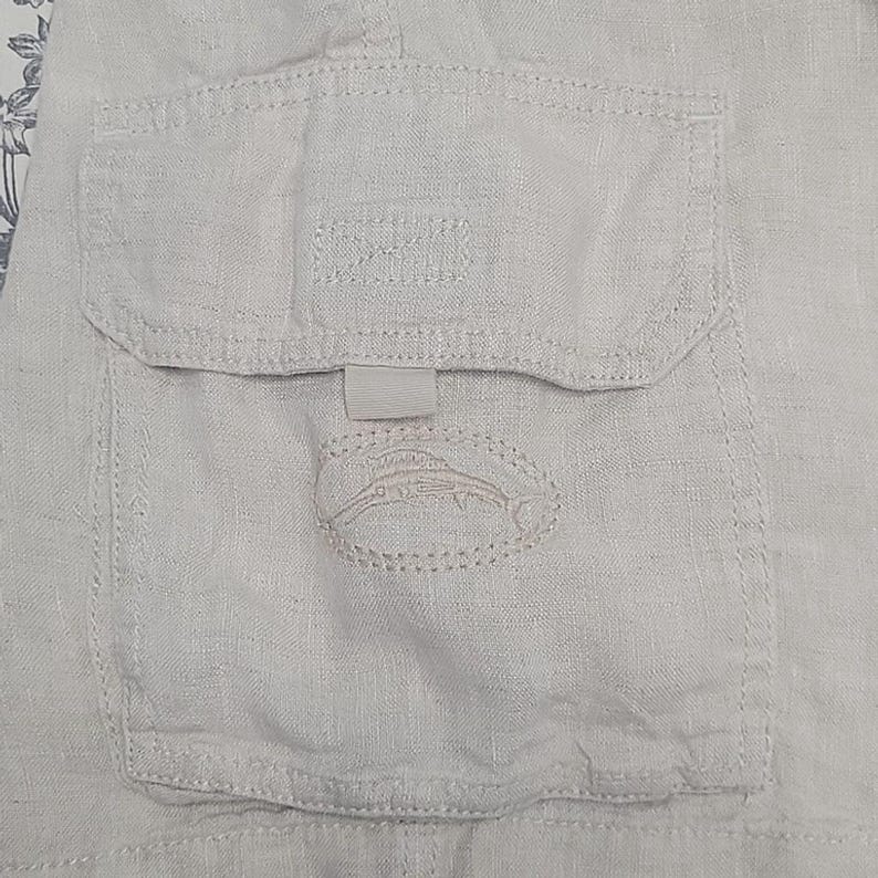 May include: Close-up of a light beige linen fabric with a pocket. The pocket has a flap and a small, stitched emblem of a fish. The fabric has visible texture and stitching details.
