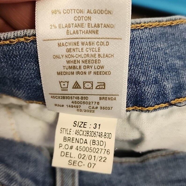 May include: A white tag with text detailing fabric composition, care instructions, and product information. The tag includes the size 31, style number, and other details. The tag is attached to a pair of blue jeans.